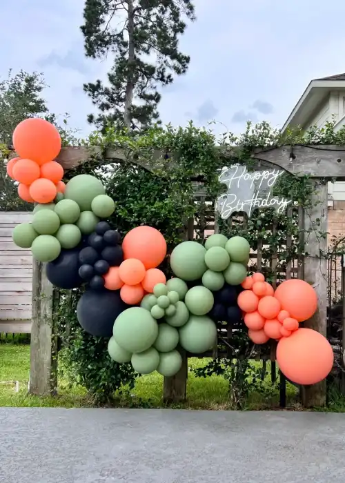 An outdoor balloon garland in coral, sage green and black along a wooden pergola, with a Happy Birthday neon sign