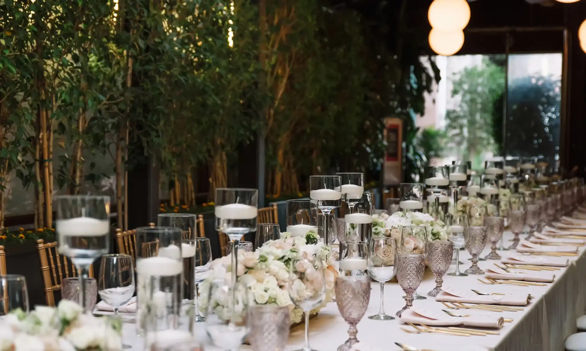 A long banquet table set with floating candles, low floral centerpieces and gold chairs under warm lighting