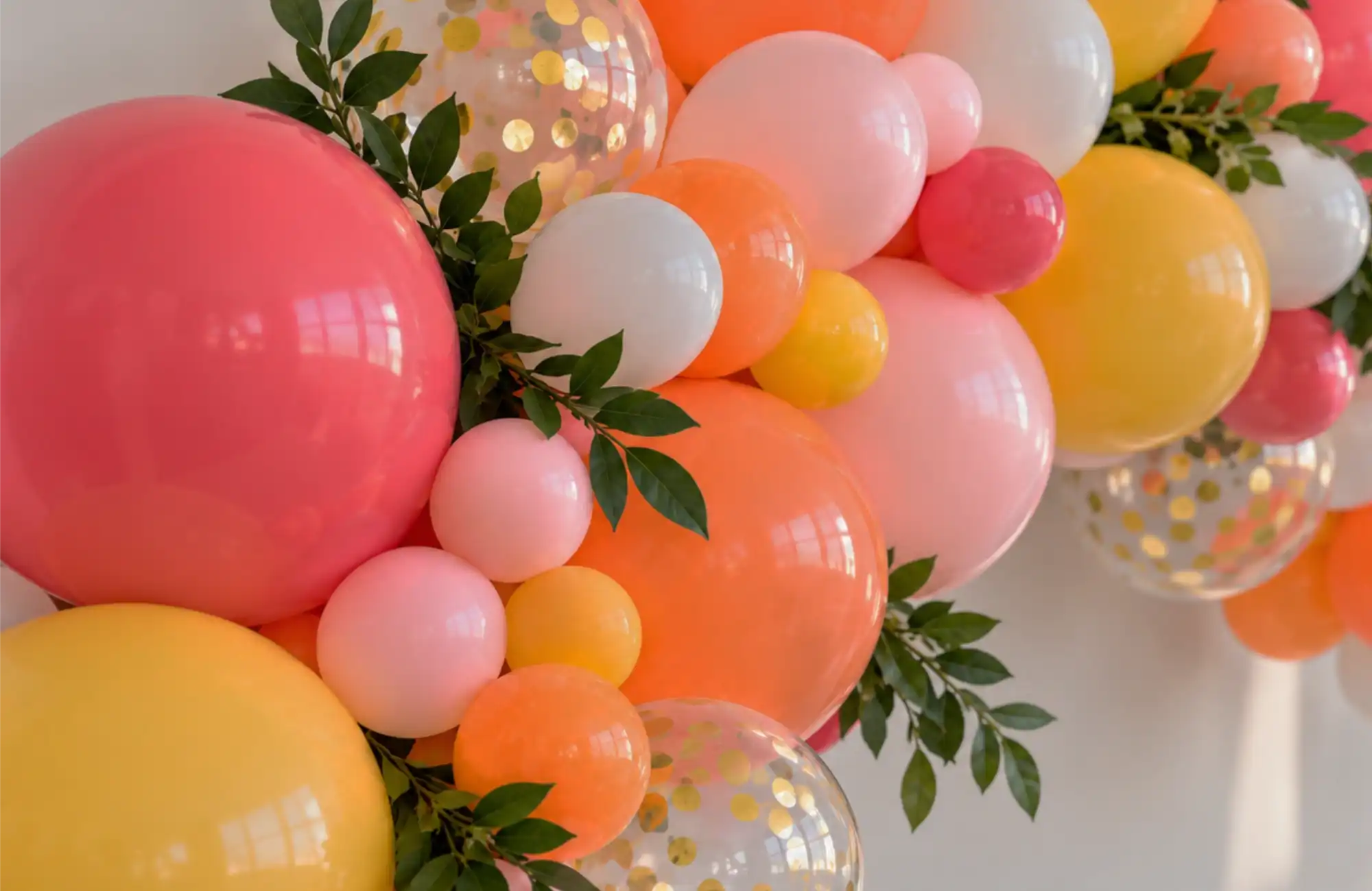 A close-up of a coral, orange, yellow and gold confetti balloon garland with greenery accents