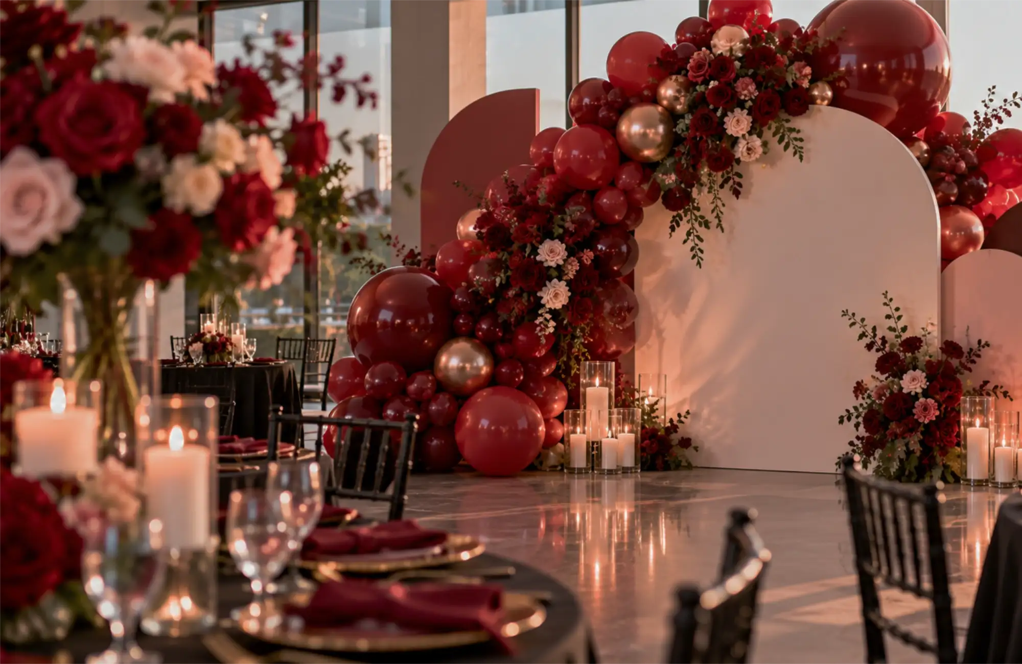 A white arch backdrop framed by a deep red balloon garland, roses and candles at a reception